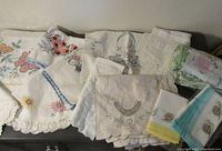 Various embroidered linens including tablecloths and napkins with pastel colors and crochet edging.