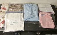 Photo showing white, pink, light blue linens packaged, some with handwriting labels indicating sizes and colors, arranged on a dark surface.
