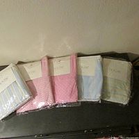Photo shows six packages of brand new crib linens: three sheets and three dusters with pastel patterns in original packaging