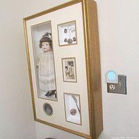 Shadow box angled on wall showing antique doll in glass compartment on left side and grouped sterling mementos, photos on right side.