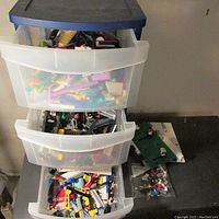 Three drawers of a plastic storage bin filled with assorted LEGO bricks and parts, with instruction books visible in the top drawer