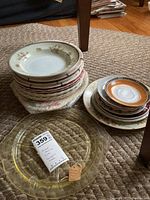 Photo showing a stack of assorted ceramic plates and saucers alongside a clear yellow Anchor Hocking post depression glass platter with scalloped edges.