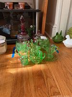 Photo showing green carnival glass tea cups, saucers, and pourers along with red decanter and jar grouped together on wooden floor.