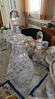 Photo shows two crystal decanters on a table with other china pieces, large decanter with diamond pattern and faceted stopper, smaller decanter behind.