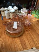 Full view of various pink Depression glass items including bowls, dessert plates, teacups with saucers, and a large platter on a wooden table with other miscellaneous ceramics in the background.