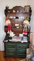 Front view of dark brown upper hutch with shelves displaying small decorative items, two stuffed dolls sitting on lower portion, and green lower cabinet with doors and scratches.