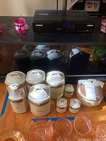 Photo showing the full lot of lusterware kitchen jars with lids arranged on a wooden floor in front of a TV. Includes various sized jars with fruit motif and name plates.