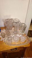 Full view of collection of clear glass and crystal vases with etched and pressed patterns arranged on wooden surface with lace doily under some items.
