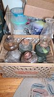 Four vintage oil lamps with clear and green glass, copper burners, and tall hurricane glass chimneys placed inside a crate on the floor surrounded by packing materials.