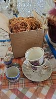 Box containing packaging material alongside small blue patterned cup, a shaker, and stacked lusterware teacups on a saucer.