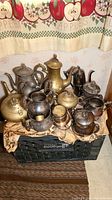 Grouped collection of teapots, creamers, and sugar urns made of pewter, silver plate and copper clustered atop a surface in front of a kitchen curtain.