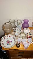 Wide view showing Nippon and hand painted teapots, milk glass mugs, lidded dishes, glass buckets and vases, Bavaria Schumann cup and bowl, and vintage wood barrel grinder on wooden surface.