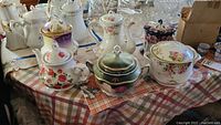 Collection of decorative teapots and covered urns on a plaid tablecloth showing various floral and gilded designs.