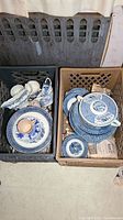 Two milk crates filled with various blue and white ironstone transferware dishes, cups, bowls, and serving pieces packed with newspaper