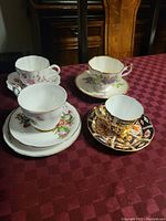 Four vintage bone china teacups with matching saucers arranged on a tablecloth, showing different floral and decorative patterns
