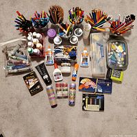 Top-view photo showing the collection of art supplies arranged on a carpet including metal containers with colored pencils and markers, bottles of paint, glue sticks, scissors, washi tape rolls, small containers with miscellaneous items, and various boxes of art tools.