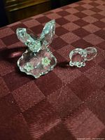 Two clear crystal figurines on a burgundy checkered cloth: hummingbird on floral base and small elephant.