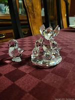 Photo of two crystal figurines on a maroon checkered tablecloth, showing a mama bird feeding three baby ducks and a smaller single baby duck figurine.
