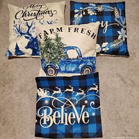 Four blue and white themed Christmas cushions arranged on carpet, featuring text and festive graphics including reindeer, vintage truck with Christmas tree, and 'Believe' message.