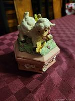 Porcelain lamb figurine on a pink hexagonal base with floral and butterfly details, shown from the side angle on a red tablecloth.