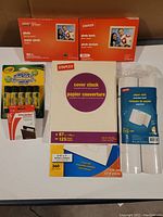 Photo of Staples photo paper packs, Staples cover stock paper, glue sticks, and thermal paper rolls displayed on a white surface.