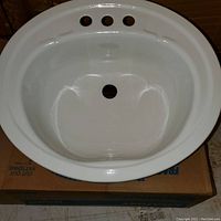 White porcelain steel oval sink with three faucet holes, shown resting on cardboard box.