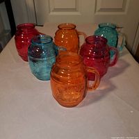 Six colored glass Mason jar mugs arranged on a white surface, in red, orange, and blue hues, showing barrel shape, handles, and embossed text