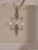 Close-up view of the clear crystal star-shaped ornament hanging by a white ribbon, highlighting its facets and shape.