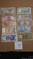 Photo showing assorted paper currency notes from various countries laid flat on a brown cardboard surface, including Venezuelan, Argentine, Chinese, Peruvian, and Mexican bills.