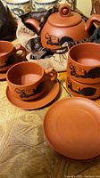 Close-up photo of reddish-brown ceramic teapot with matching coffee cups, saucers, and fabric pouch bag.