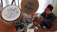 Photo showing vintage blue and white stoneware plates in round packaging box, Currier & Ives style bell, vintage doll, salt and pepper shakers, and additional decorative items arranged on a wooden table.