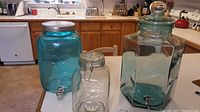 Three glass containers on kitchen counter: one large clear hexagonal dispenser with lid and spigot, one tall aqua glass dispenser, and one smaller clear glass jar with clamp lid.