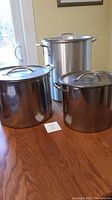Front view of three large polished stainless steel pots with lids on a wooden table, showing their height and handles.