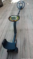 Full view of the metal detector showing the handle, armrest, shaft, and circular search coil on wooden deck floor.