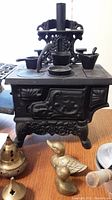 Miniature cast iron stove with pots visible, two duck figurines in front, and brass decorative item with perforations.