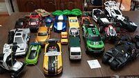 Photo showing 14 various remote control and battery operated cars on a table with mixed colors and styles including racing cars, pickup trucks, and specialty vehicles.