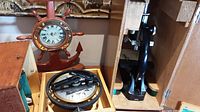 Photo showing the black vintage microscope on the right side and the nautical compass in wooden case at the center, with a ship wheel themed clock behind them.