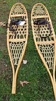 Full view of the pair of vintage wooden snow shoes with woven webbing and leather straps on grass.