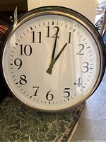 Front view of the IKEA round wall clock with white face and black numerals and hands on a green marble-like surface.