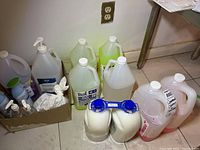 Wide view showing multiple large jugs and smaller bottles of cleaning supplies and sanitizer on floor near wall outlet.