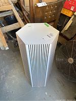 Front and side view of the white laminate six-sided pedestal with vertical black stripes, placed on a concrete floor next to a fan and wooden furniture.