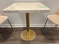 Front view of square dining table with white stone top and gold metal base, between two chairs.