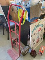 Full view of red metal hand truck dolly with large wheels, standing upright on concrete floor surrounded by various items.