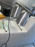 Two stainless steel heating lamps assembled on a metal stand with an electrical cord visible, showing the full frame and lamps in a kitchen or commercial setting.