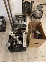 Photo showing plastic crates and a cardboard box with various kitchen utensils and containers, including metal cups, pots, and ladles.