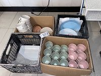 Overall view of four boxes containing pastel colored cups, plates, and bowls in a mixture of green, pink, white, blue, and brown hues on a tiled floor.
