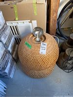 Ceiling lamp with woven rattan shade on the floor surrounded by boxes, showing top metal fixture and tag.