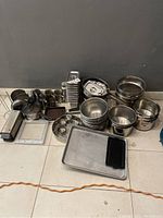 Wide shot of the lot showing stainless steel mixing bowls stacked, metal condiment containers, various pans, measuring tools, and kitchen utensils on tiled floor against a grey wall.