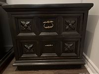 Front view of the black wood side table showing the two drawers with carved floral panels and metal handles.