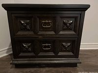 Front view showing dark finish, two drawers with carved designs and metal handles.